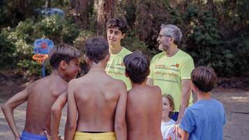 A group of people standing outdoors, some shirtless, in a natural setting with trees and a basketball hoop in the background.