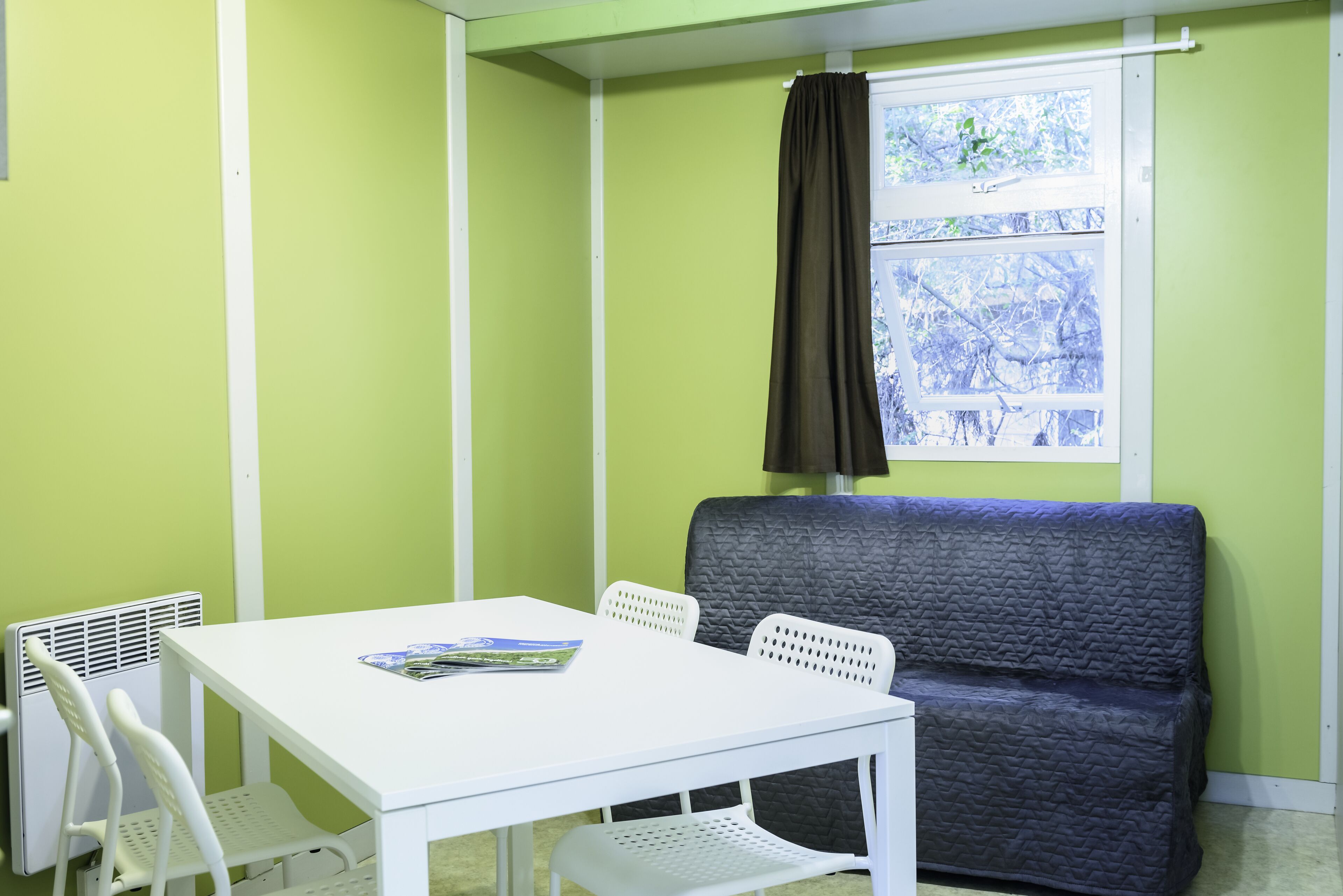Een kamer met een witte tafel, twee witte stoelen, een donkerblauwe bank en een raam met een zwart gordijn.