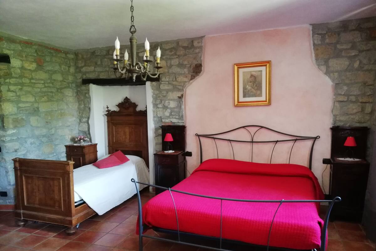 A bedroom with a stone wall, a wooden wardrobe, a bed with a red bedspread, a chandelier, and two bedside lamps.