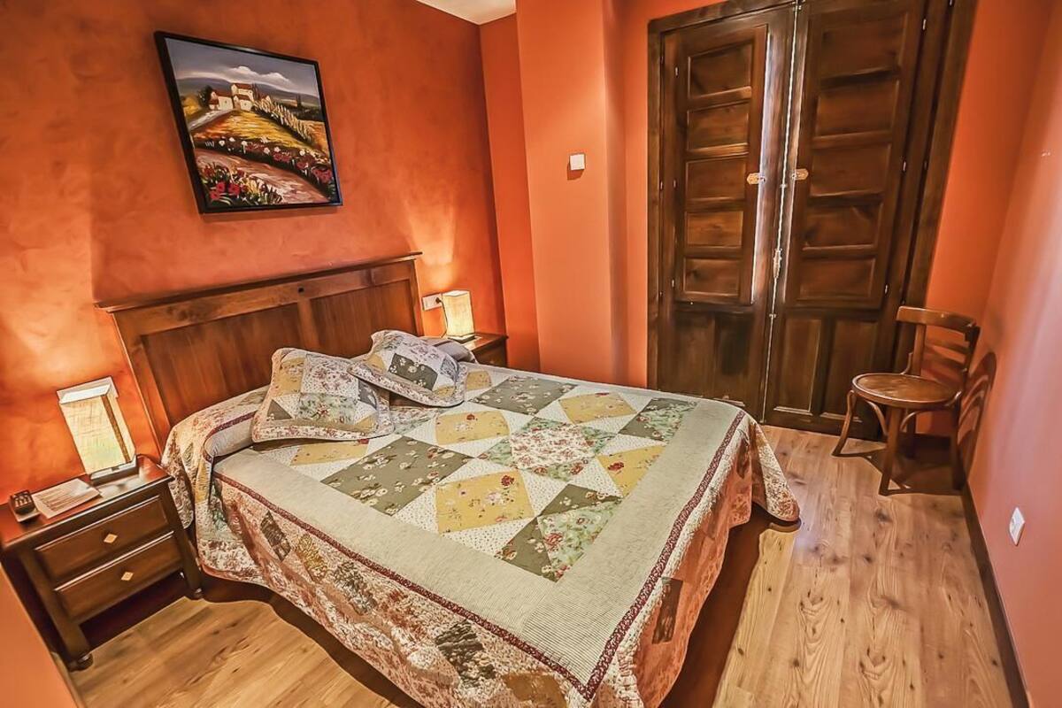 A bedroom with a bed, nightstand, lamp, wooden wardrobe, and a painting on the wall.
