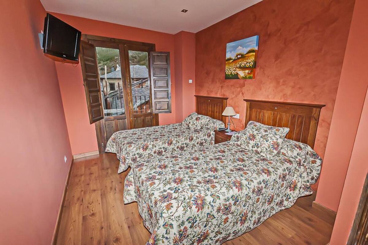 A bedroom with a bed, a TV mounted on the wall, a painting, and a window with a view of a house.
