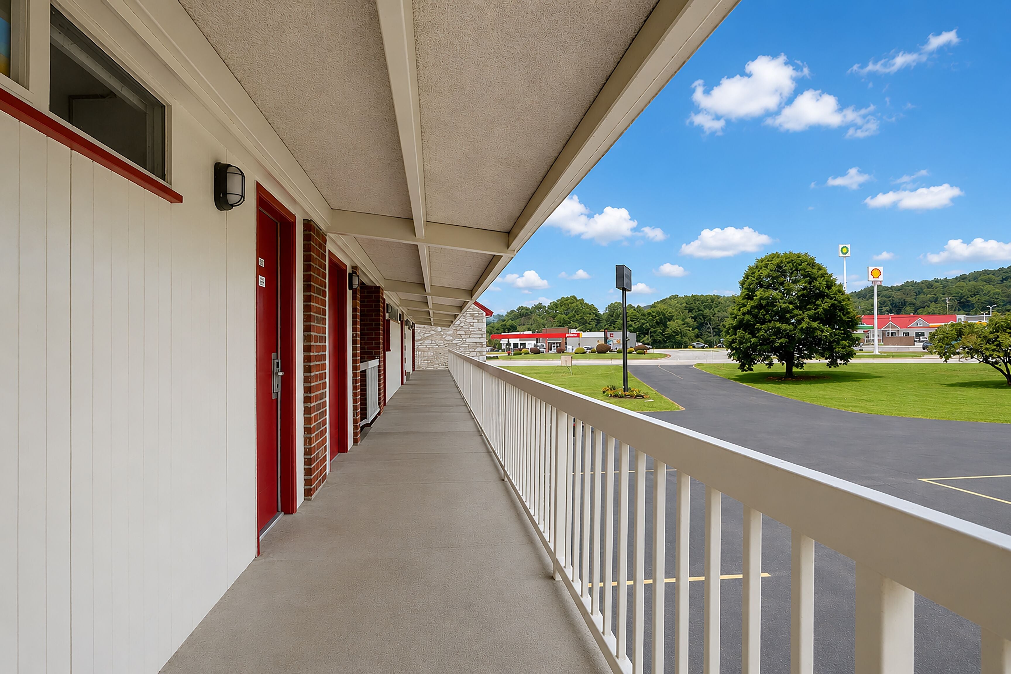 A motel corridor with a covered walkway, red doors, and a view of a parking lot and gas station in the distance.