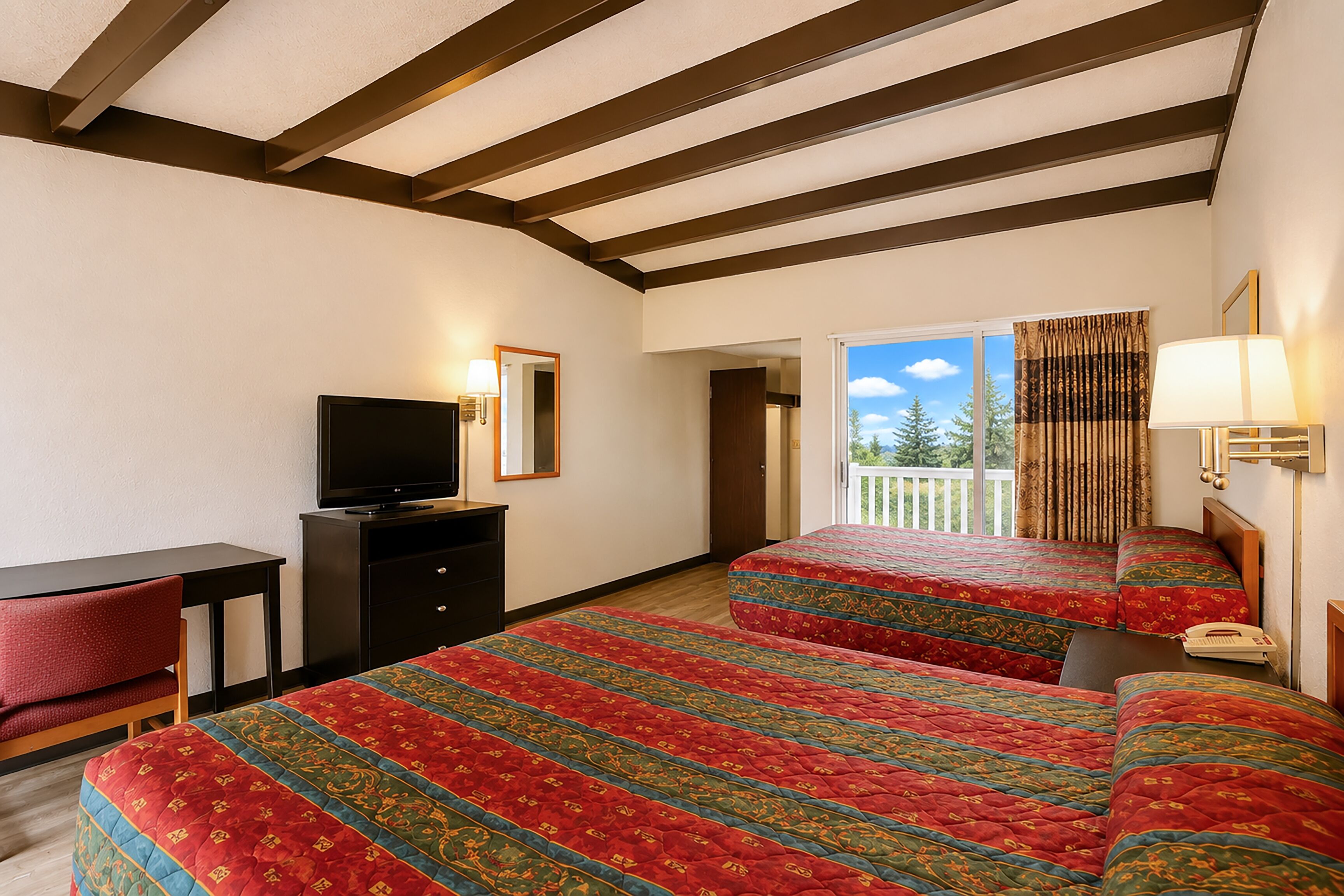 A hotel room with a bed, a desk, a television, and a balcony with a view of trees.