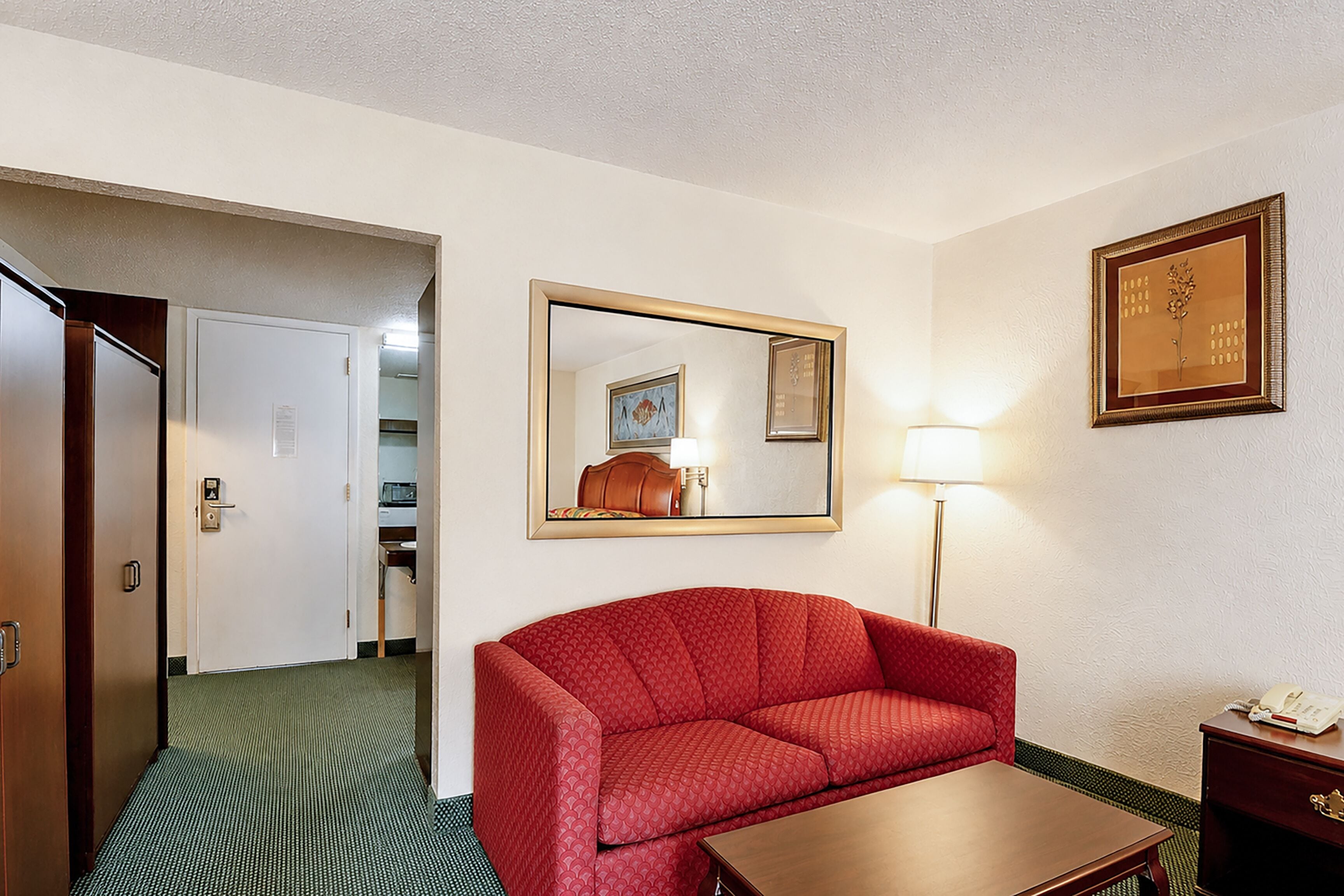 Ein Hotelzimmer mit einem roten Sofa, einem Holznachttisch, einer Lampe, einem gerahmten Bild an der Wand und einer Tür zu einem Badezimmer.