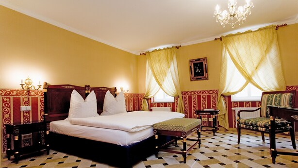 A room with a large bed, a small table, a chair, and a chandelier.