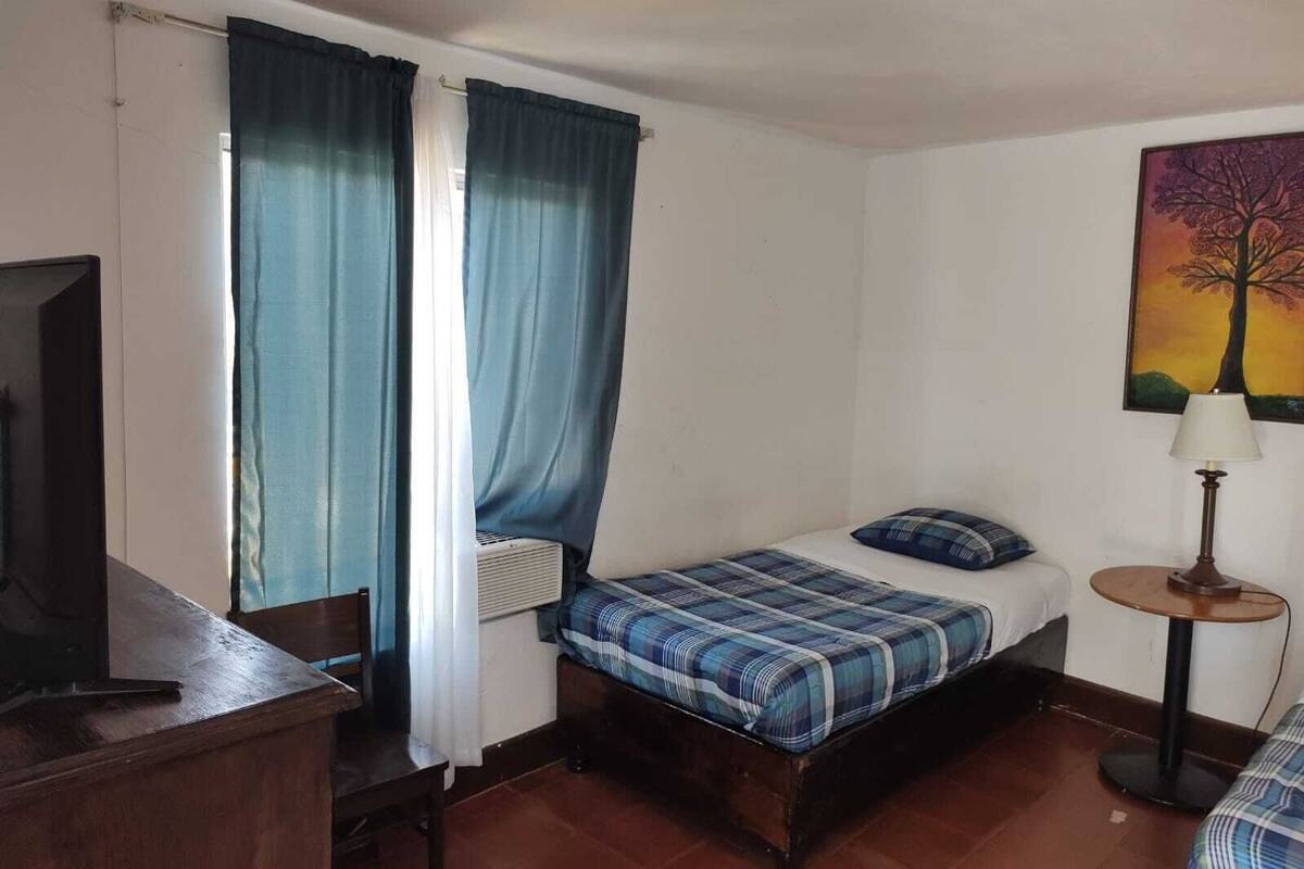 A small, simple room with a bed, a desk, and a chair. There is a window with curtains, a painting on the wall, and a lamp on a small table.