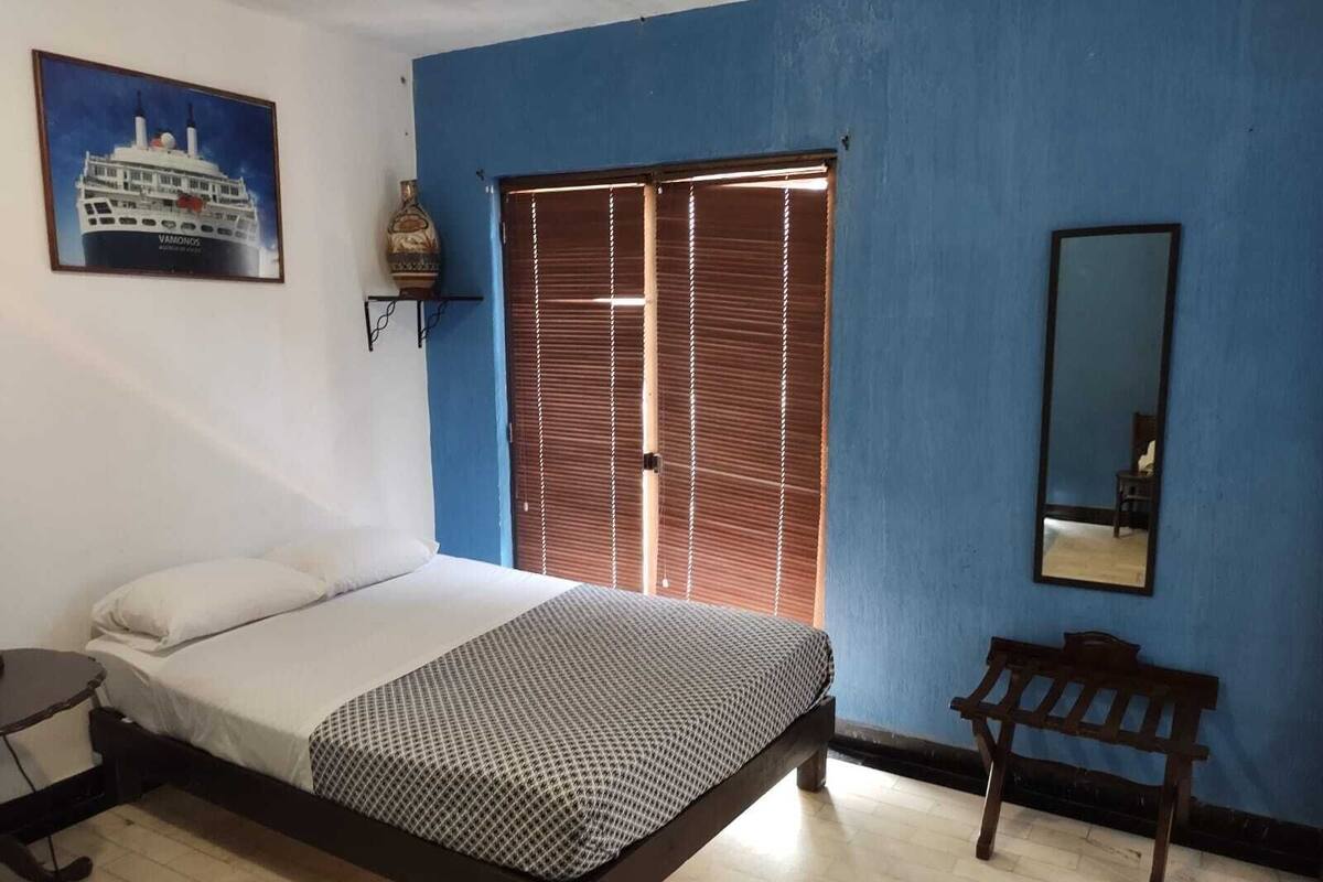 A bedroom with a bed, a desk, a chair, a window with blinds, a mirror, and a wall-mounted shelf with a vase.