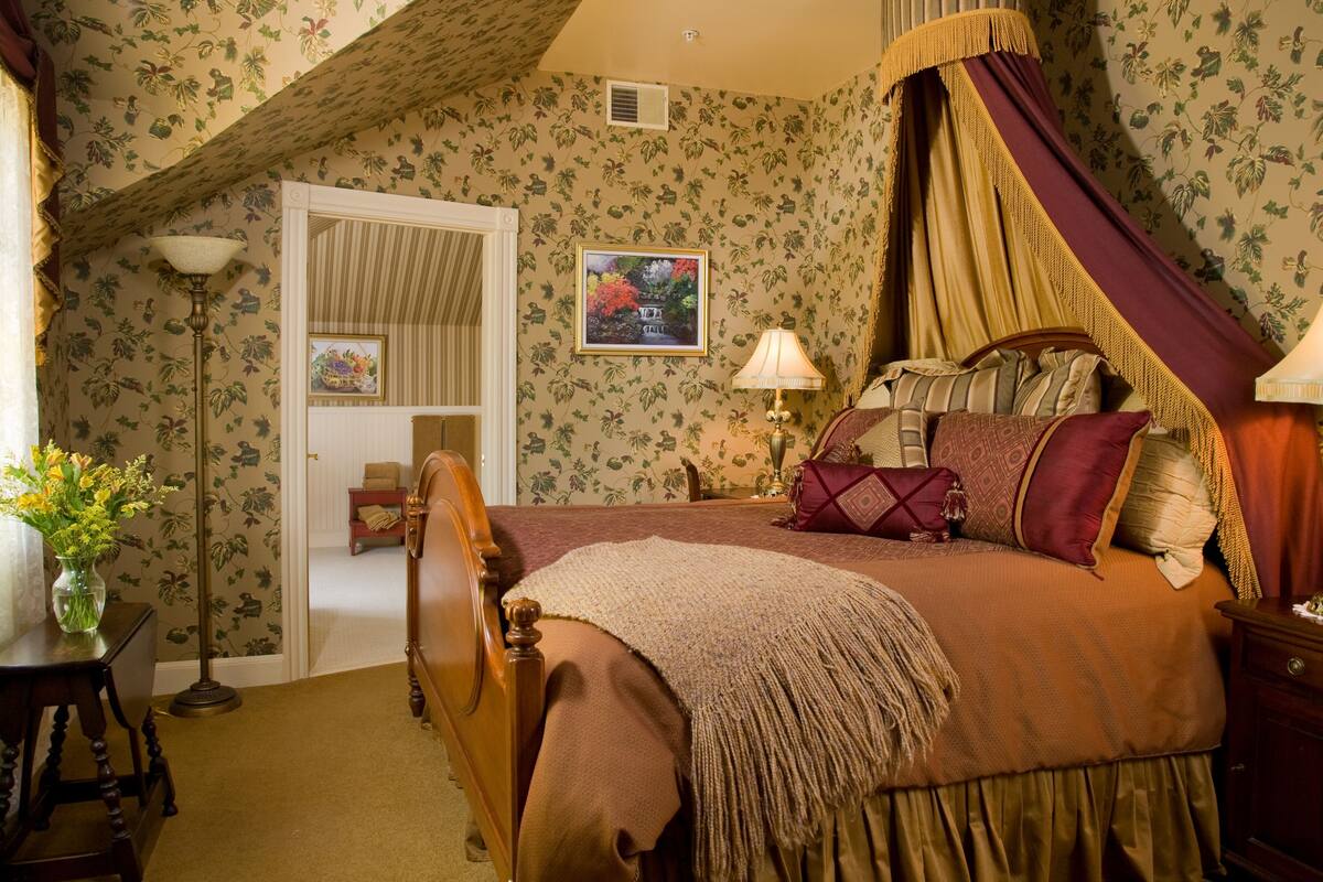 A bedroom with a four-poster bed, a nightstand with a lamp, a bedside table with a vase of flowers, and a door with a mirror and a framed picture.