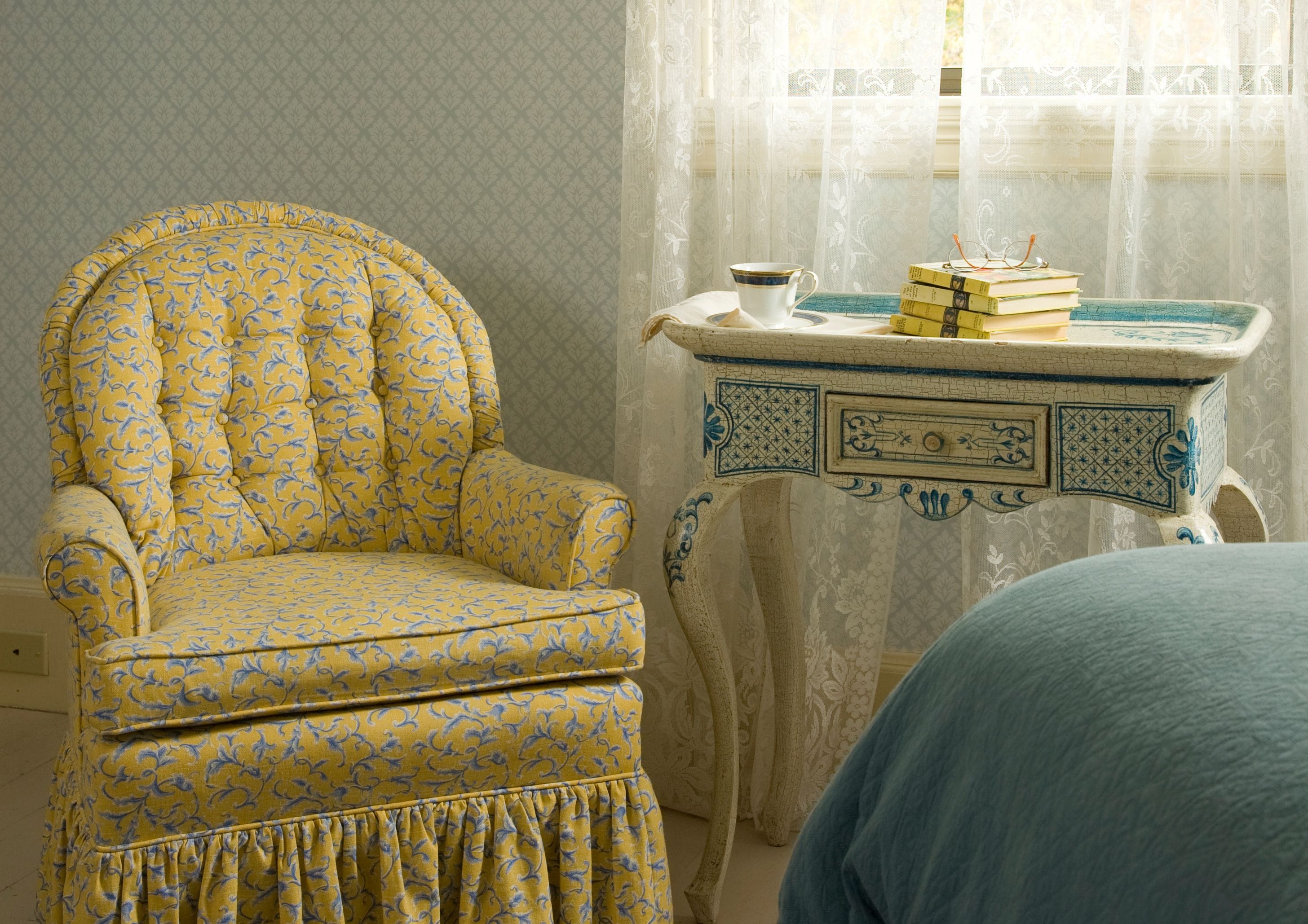 A room with a patterned armchair, a small table with books and glasses, and a sheer curtain.