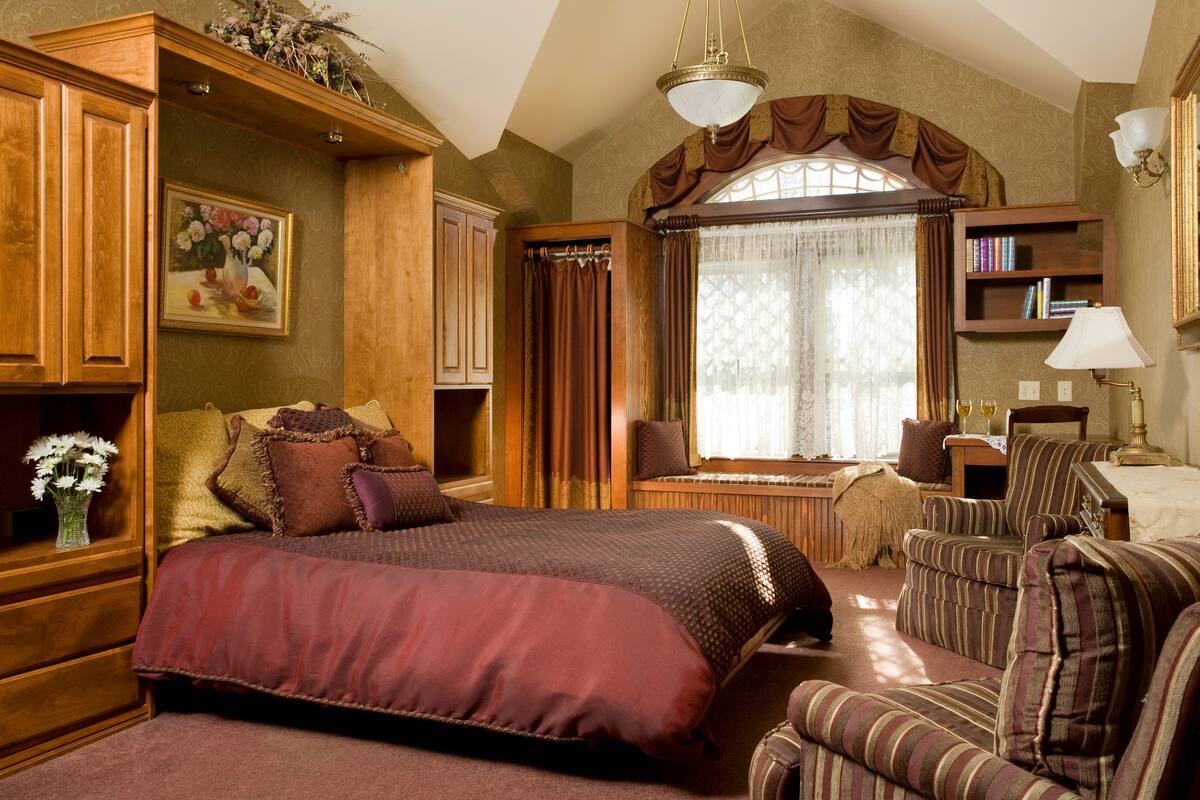 A spacious bedroom with a large bed, a seating area with a chair and a small table, wooden cabinets, and a bookshelf.