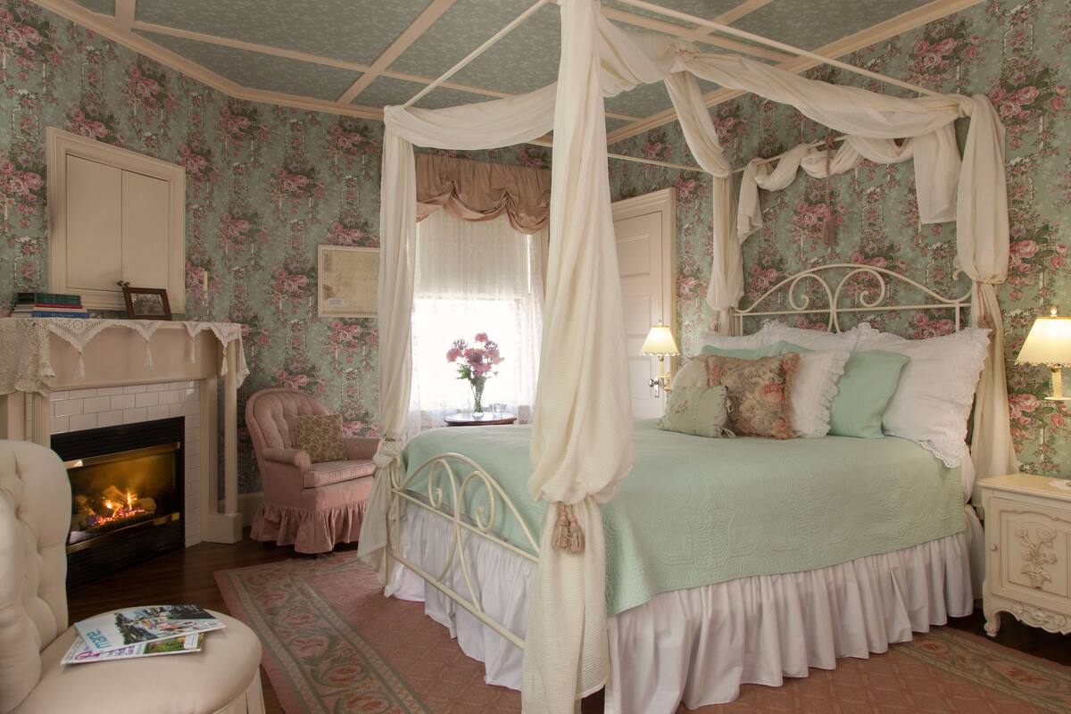 A bedroom with a four-poster bed, a fireplace, a chair, and a nightstand.