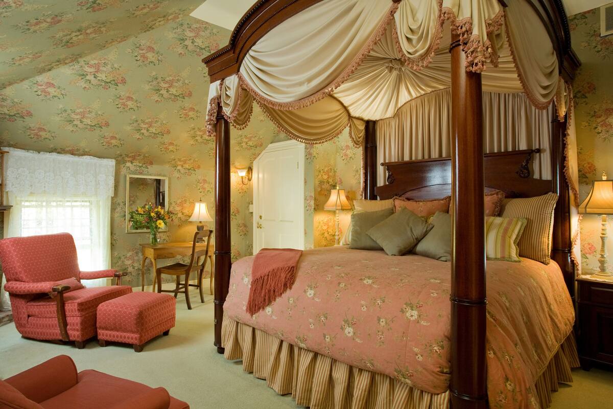 A four-poster canopy bed with floral bedding, a wooden nightstand, and a chair with a matching cushion.