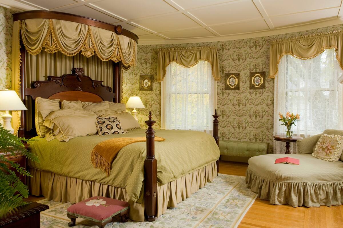 A four-poster bed with a wooden headboard, a green upholstered bench, and a small stool with a floral cushion.