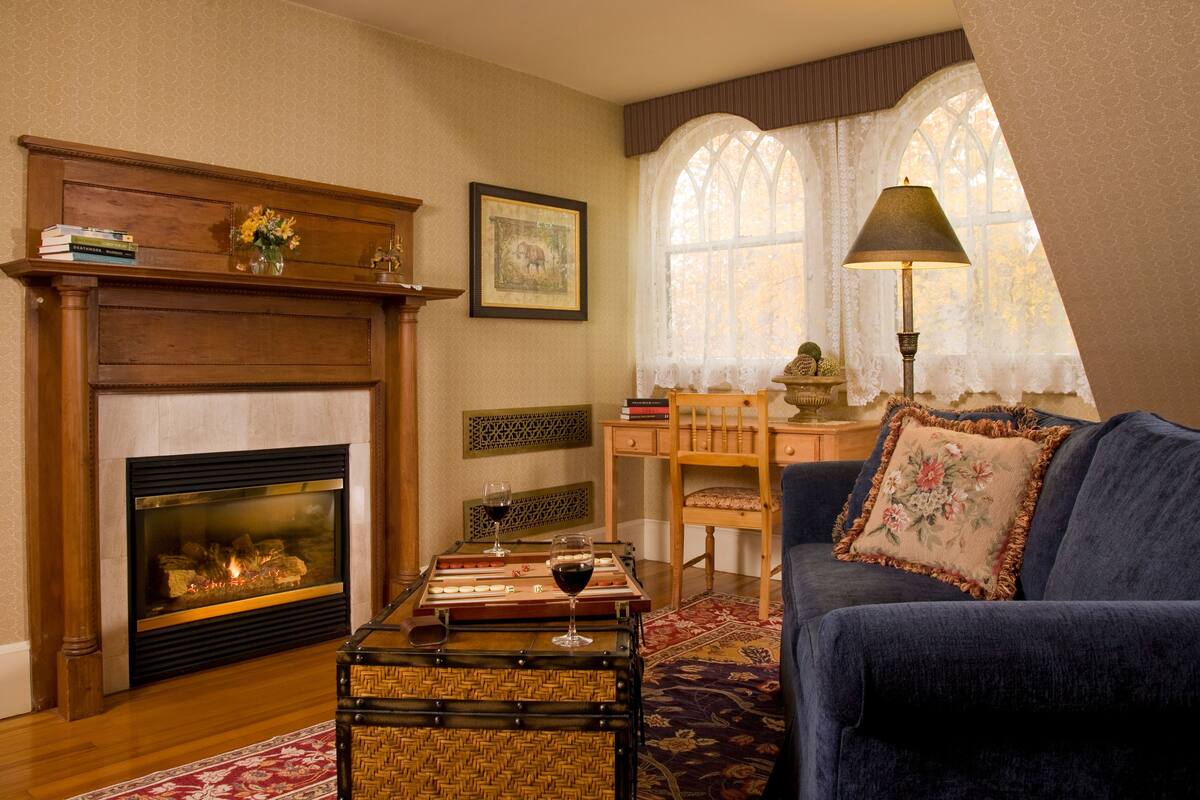 A cozy living room with a fireplace, a blue sofa, a wooden table with a chess set, and a window with lace curtains.