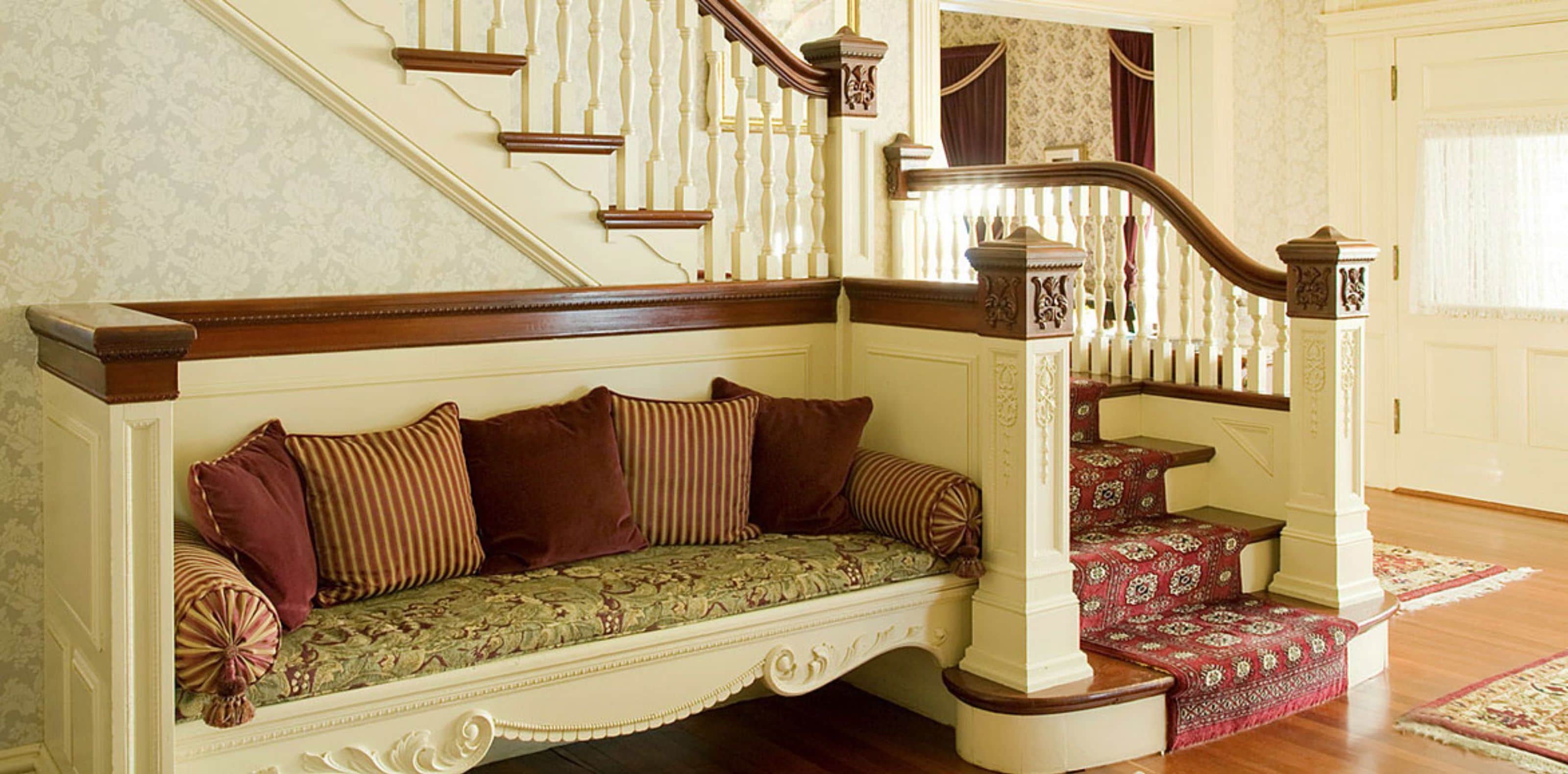 A traditional staircase with a wooden handrail and white balusters, leading to a landing with a red patterned rug.