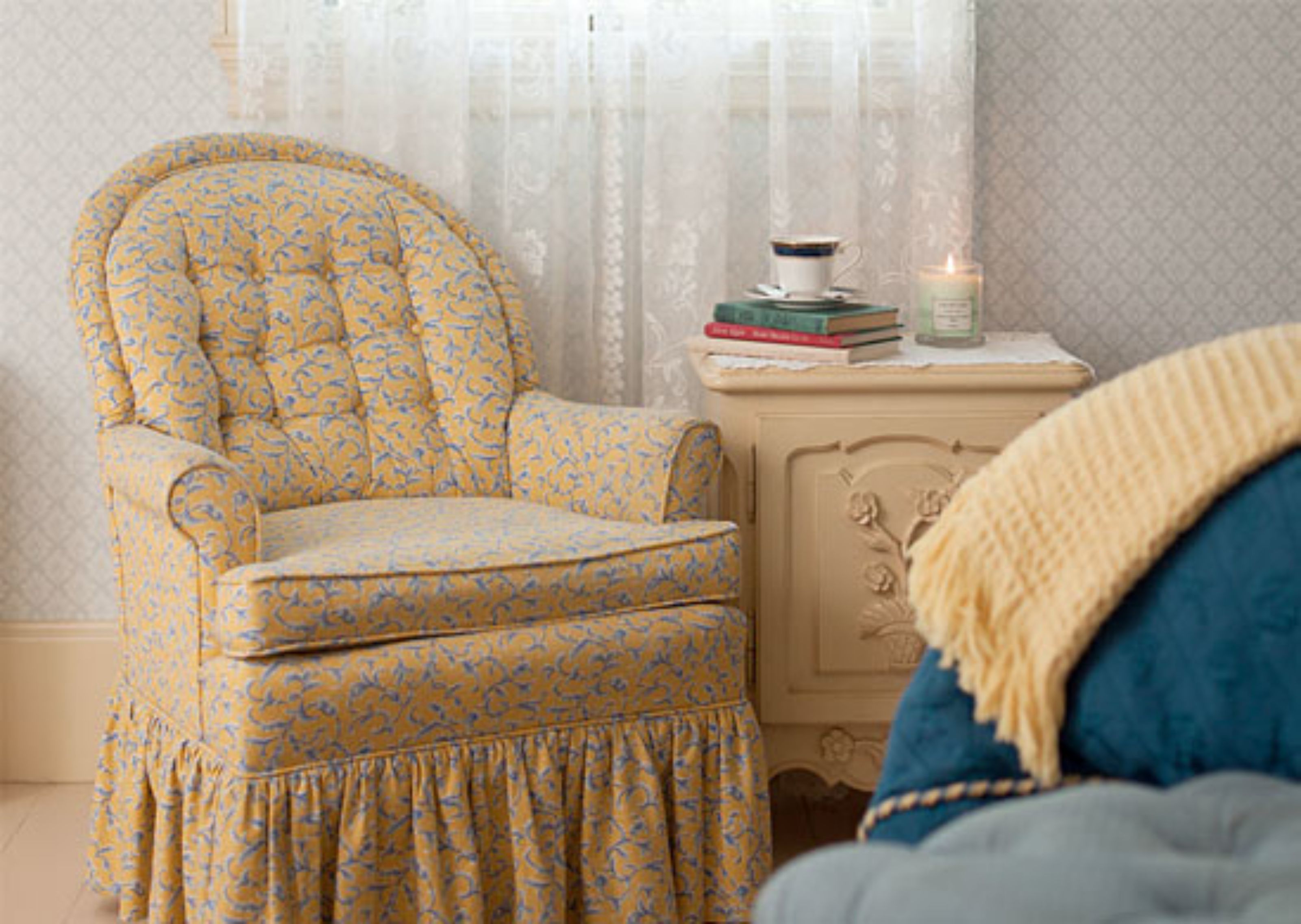 A room with a patterned armchair, a bedside table with books and a candle, and a blue upholstered headboard.