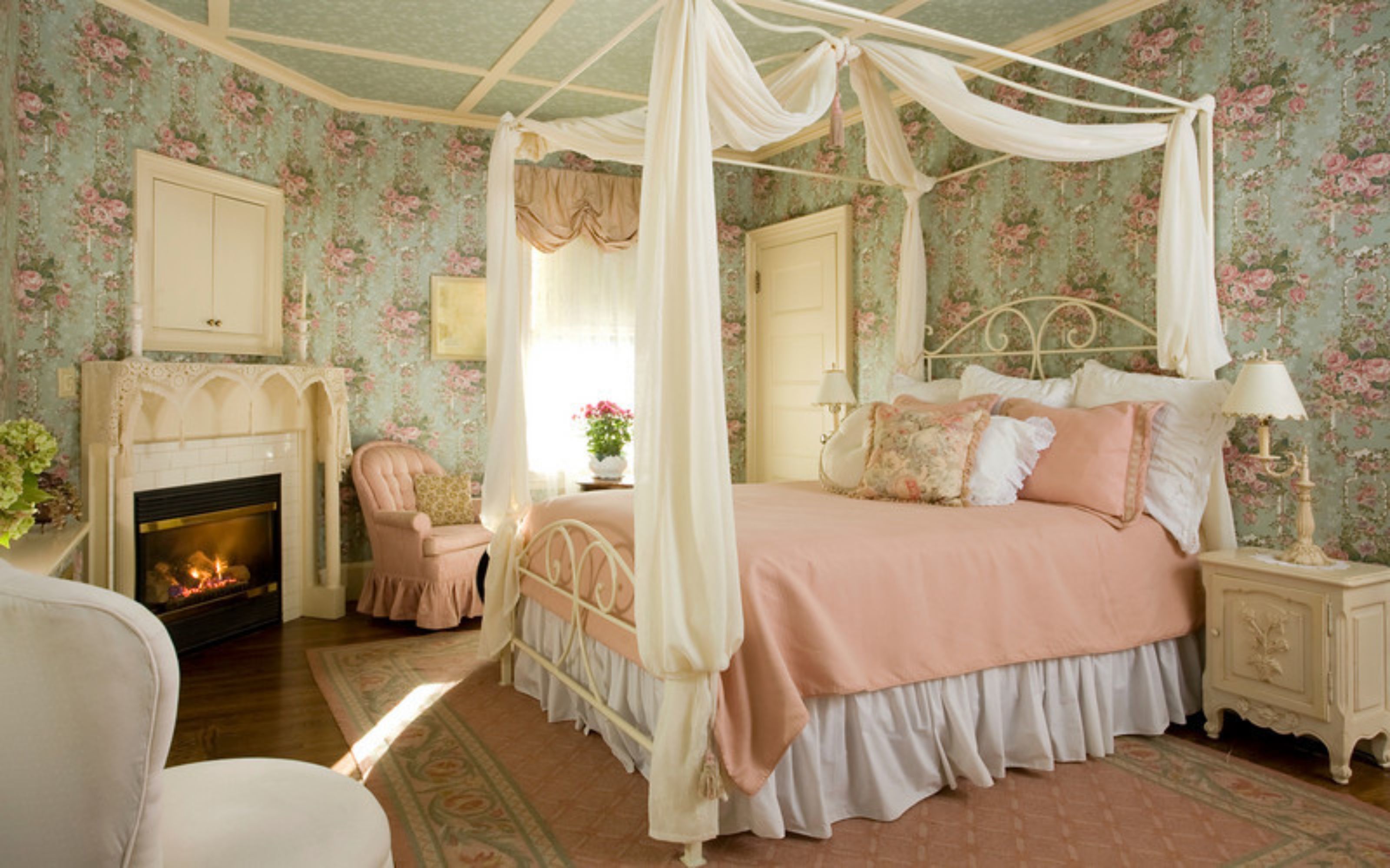 Een slaapkamer met een hemelbed, een open haard, een stoel en bloemenbehang.