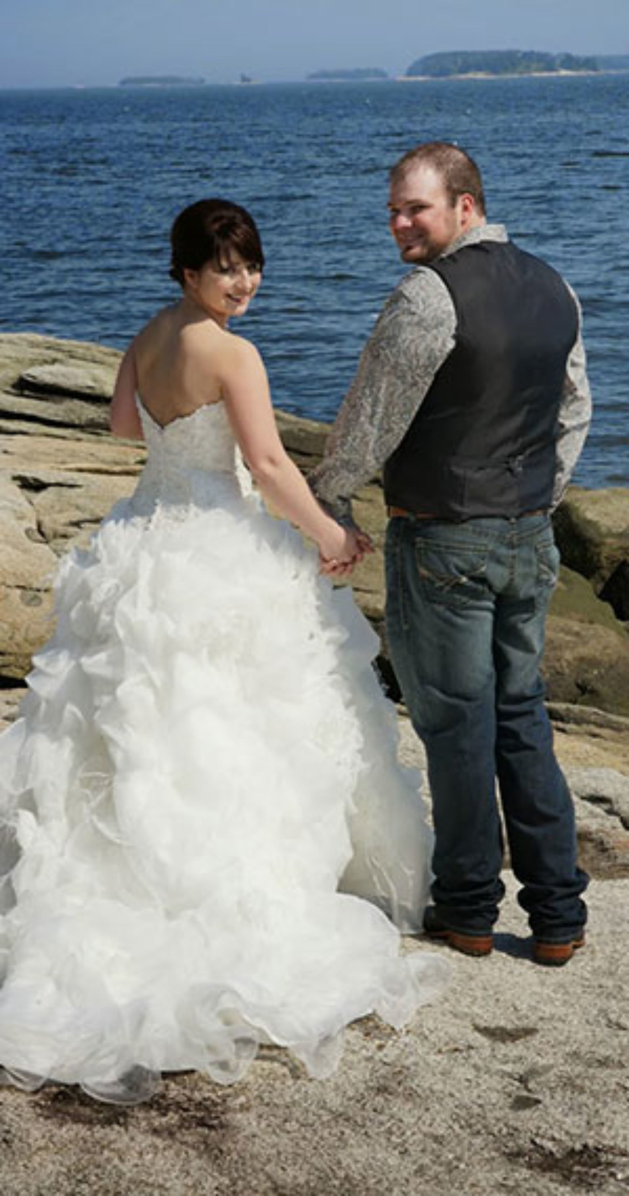 A couple holding hands on a rocky shoreline with a body of water in the background.