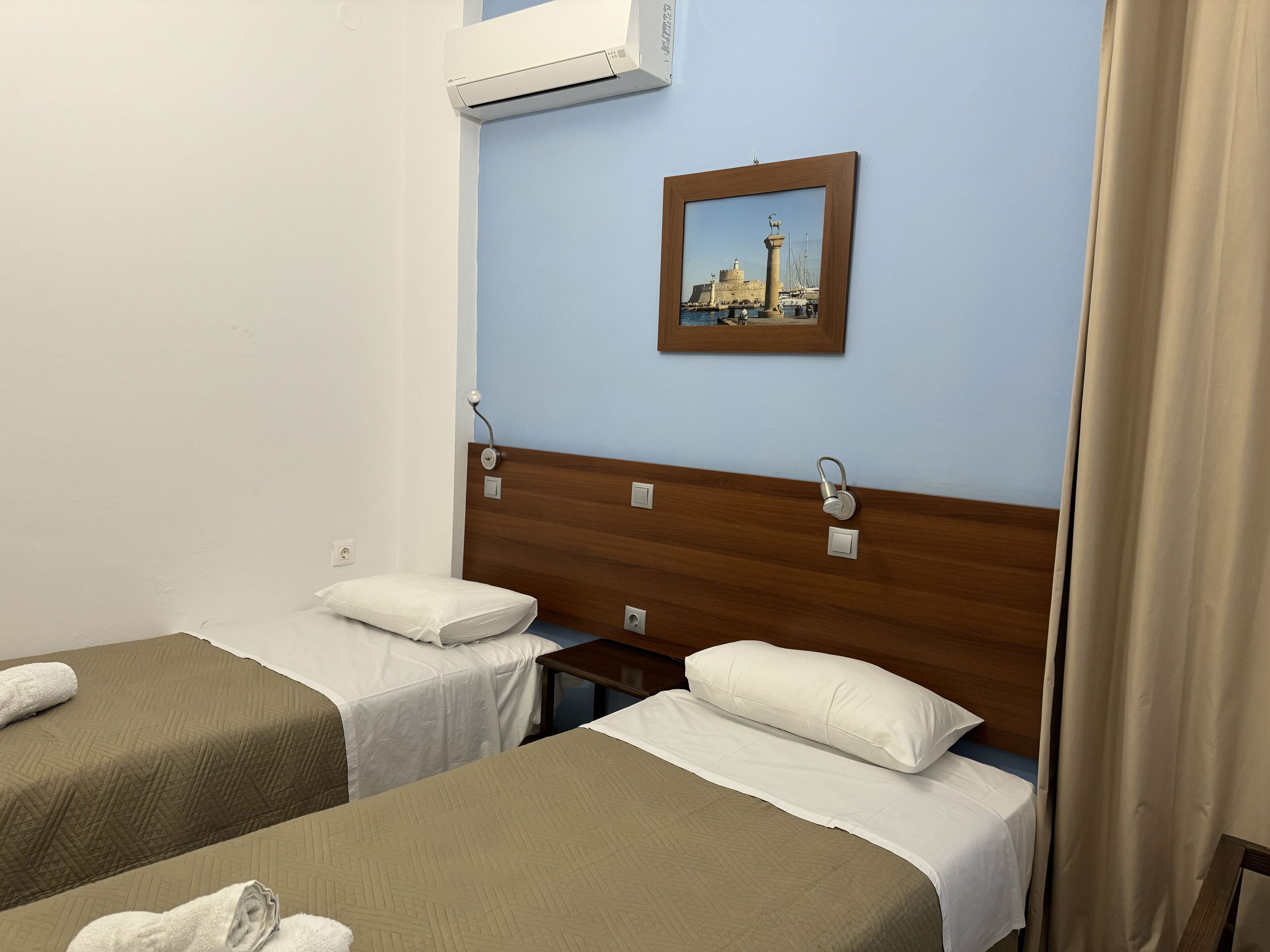 Une chambre d’hôtel avec deux lits simples, une tête de lit en bois, un tableau encadré au mur et un appareil de climatisation.