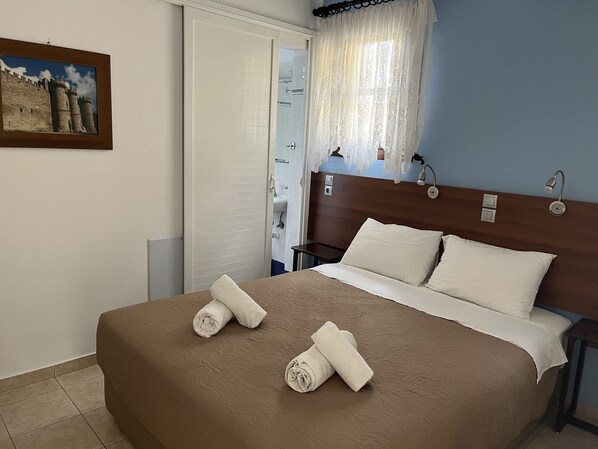 Hotelkamer met een bed, kussens, een hoofdeinde, een nachtkastje, een lamp, een schilderij aan de muur en een zichtbare badkamer door een geopende deur.