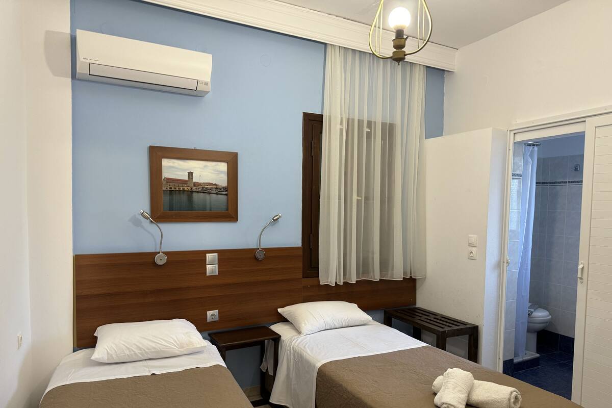 Habitación de hotel con dos camas, una mesita de noche, aire acondicionado, una ventana con cortinas translúcidas, un cuadro en la pared y un baño visible a través de una puerta abierta.