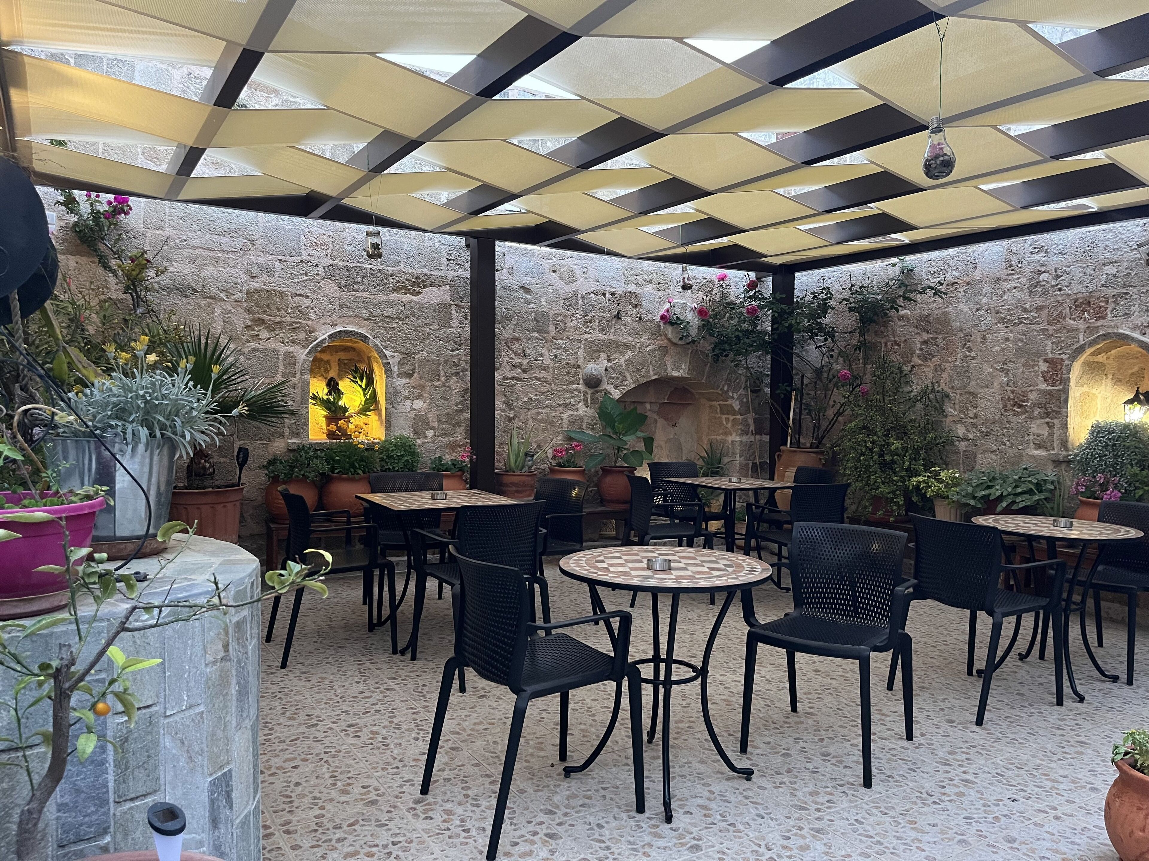 Un café en plein air avec des murs en pierre, des plantes en pot et des chaises en osier noir.