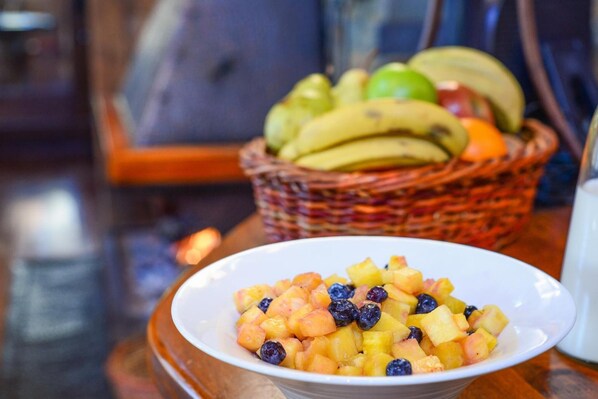 Un tazón de ensalada de frutas con una cesta de plátanos y manzanas al fondo.