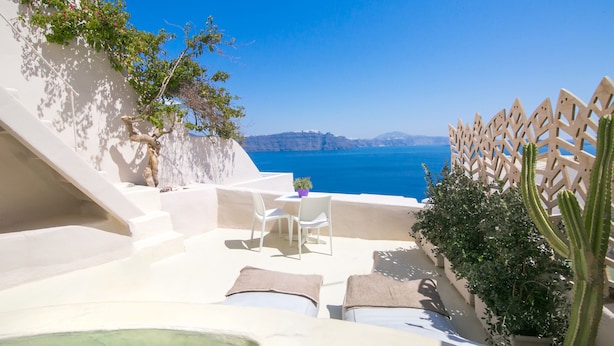 A sunlit outdoor seating area with a view of the sea, white walls, and a cactus plant. - Morfi Cave House (Santorini)