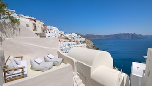 A coastal hotel with white buildings, a staircase leading to a terrace with lounge chairs, and a view of the sea and distant mountains. - Morfi Cave House (Santorini)