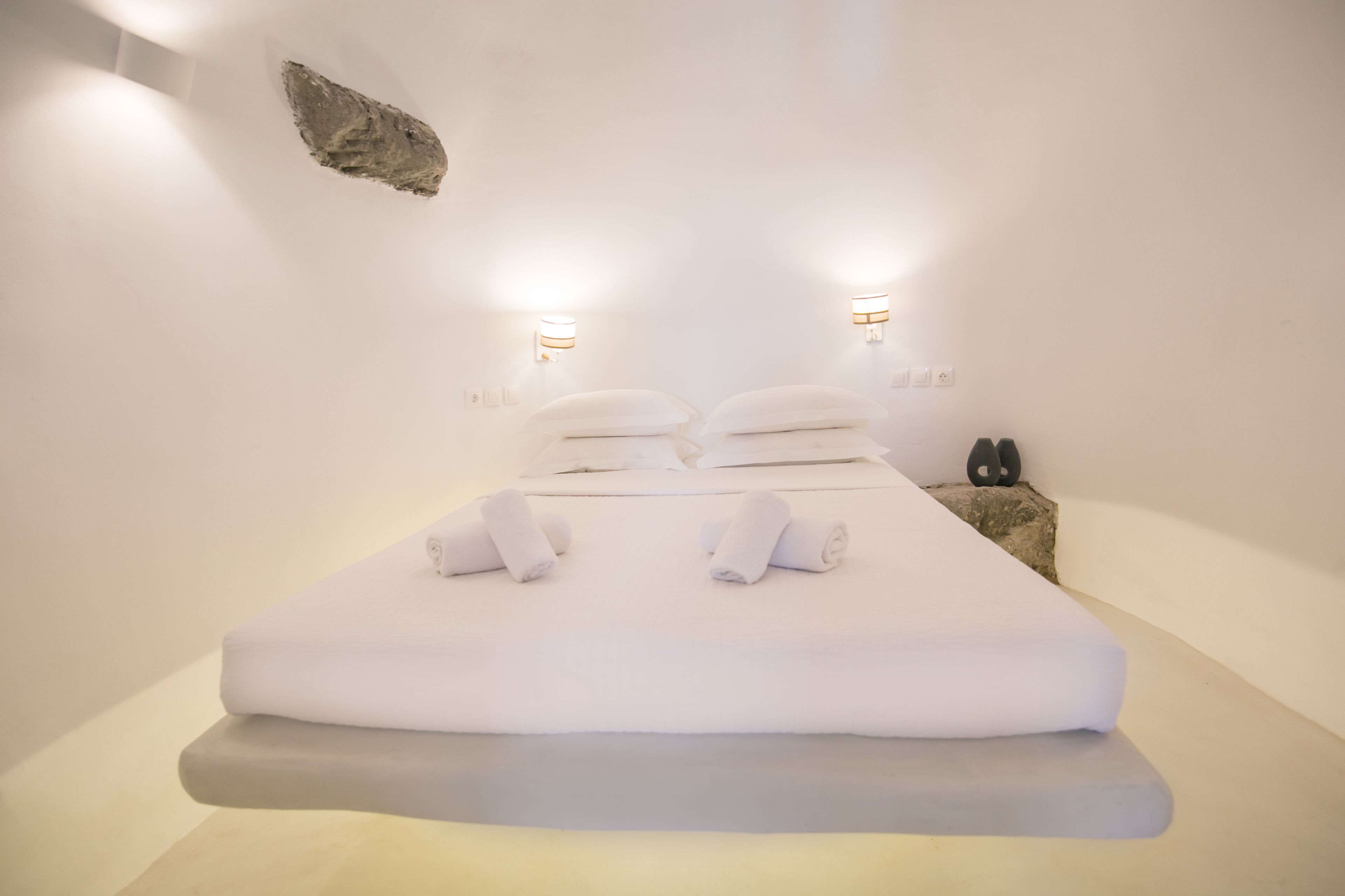 A minimalist hotel room with a large bed, two wall-mounted lights, and a small nightstand with a pair of shoes.