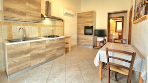 Una cocina funcional con armarios de madera, estufa a gas y horno empotrado. Cuenta con una zona de comedor con mesa y sillas, y un televisor montado en la pared.