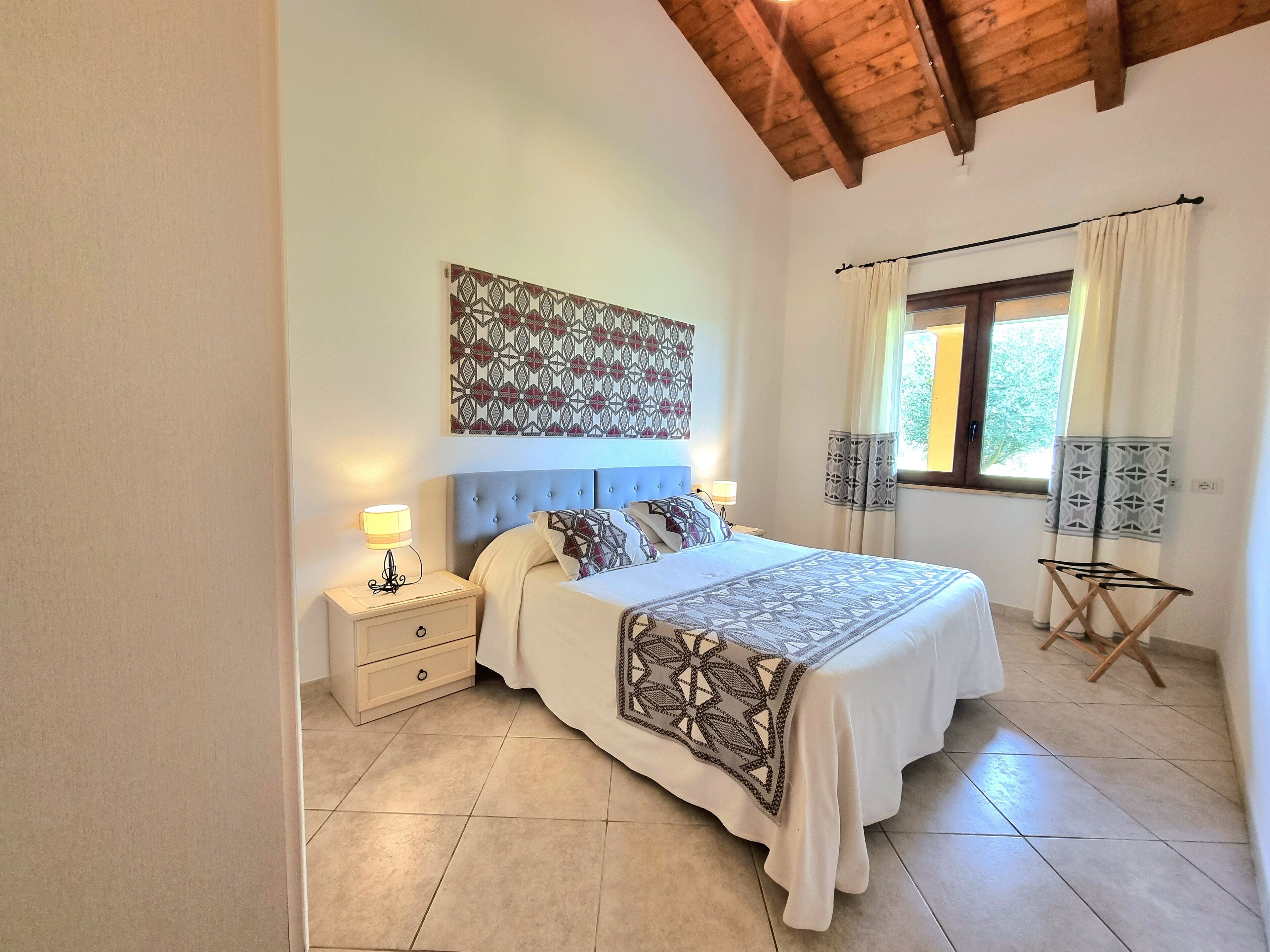 A bedroom with a bed, nightstands, a window with curtains, a ceiling fan, and a wall-mounted artwork.