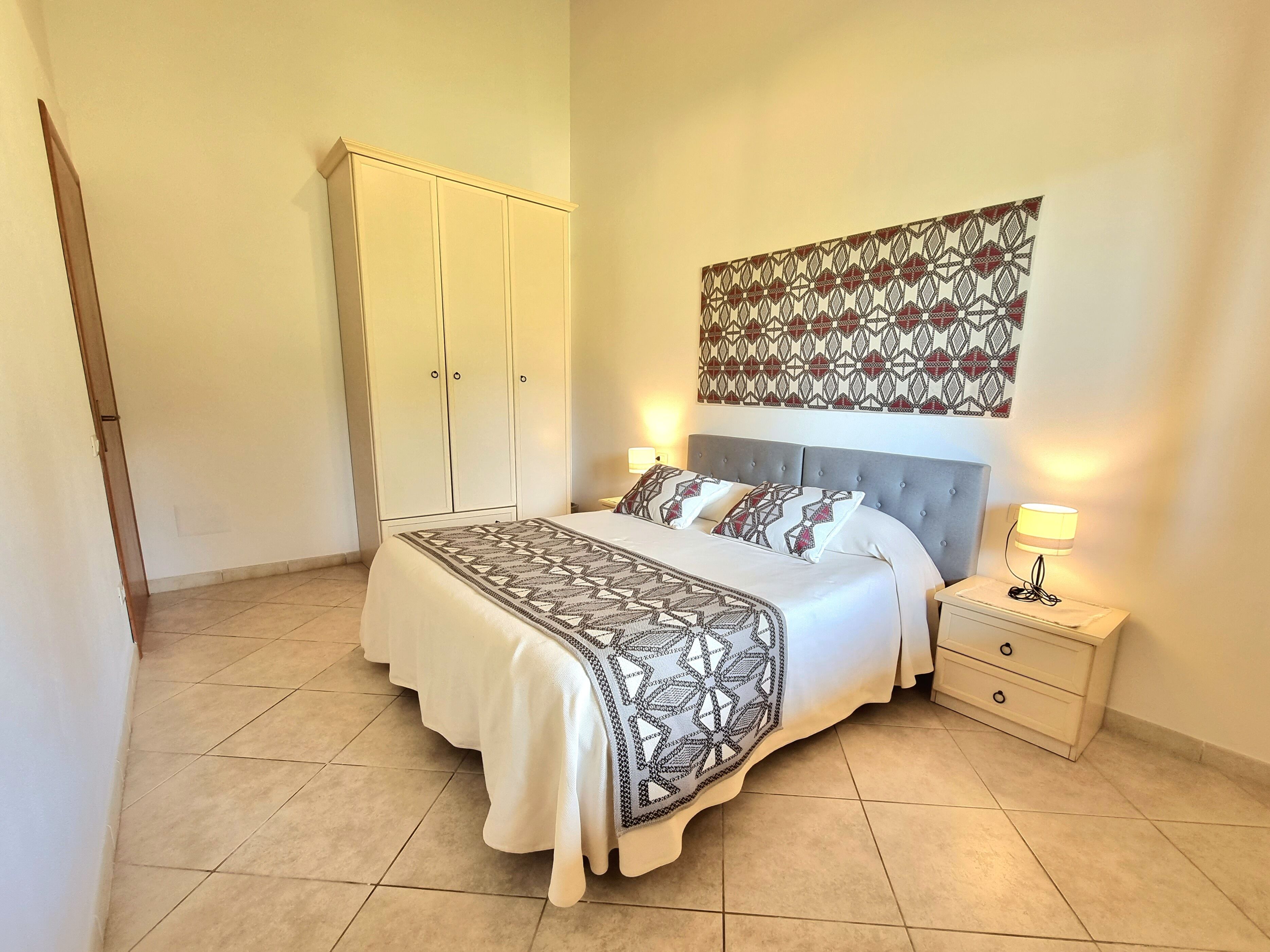 A bedroom with a bed, nightstands, a wardrobe, and a patterned wall hanging.