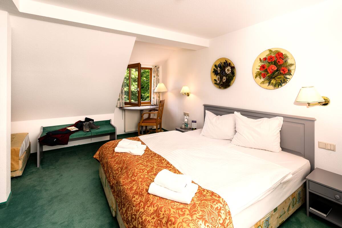 A hotel room with a large bed, a desk with a chair, a window with curtains, and two wall-mounted lamps.