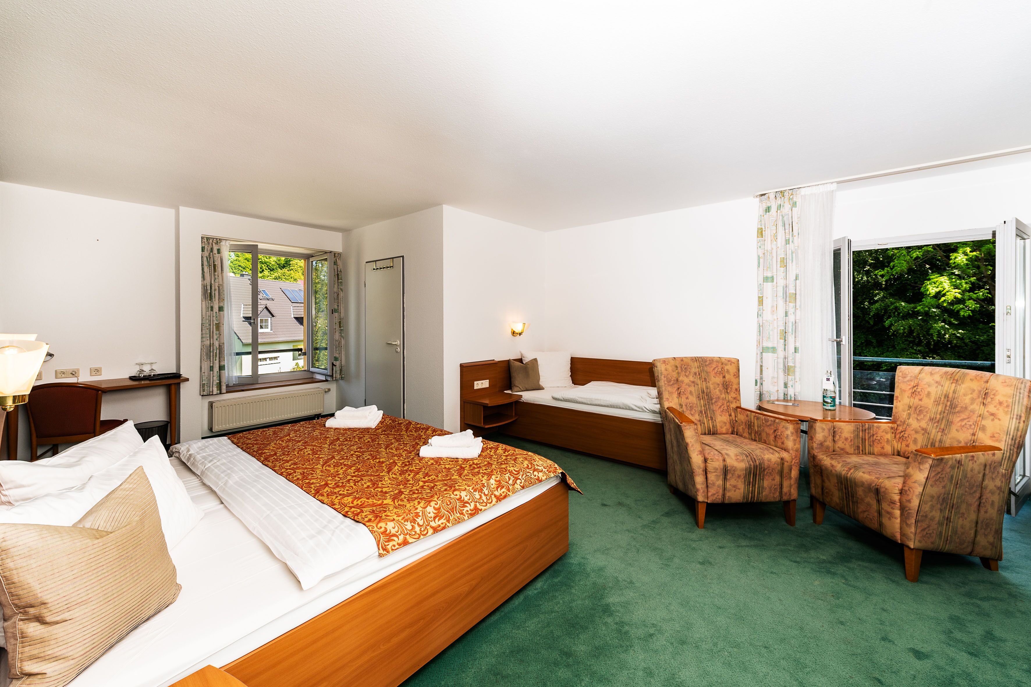 A hotel room with a large bed, a desk, two armchairs, a small table, and a window with a view of trees.