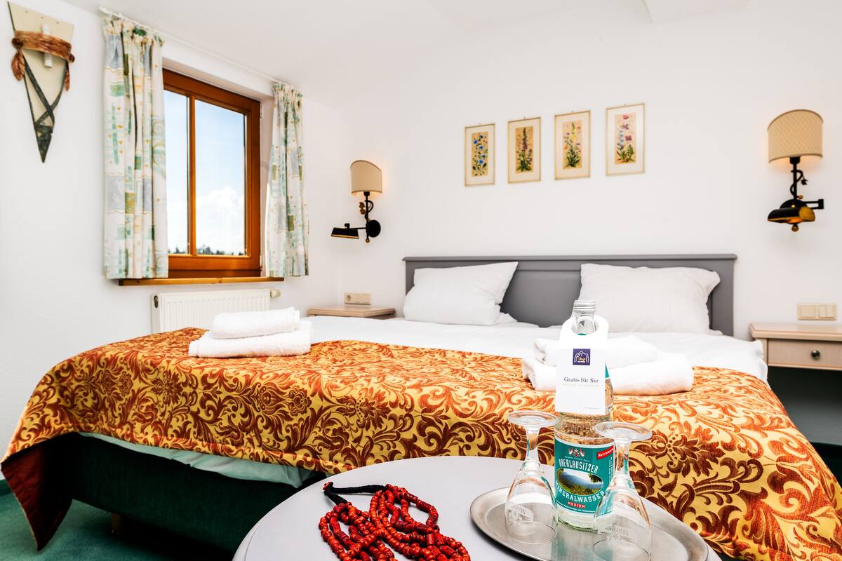 A hotel room with a large bed, a round table with a bottle and a glass, and a window with curtains.
