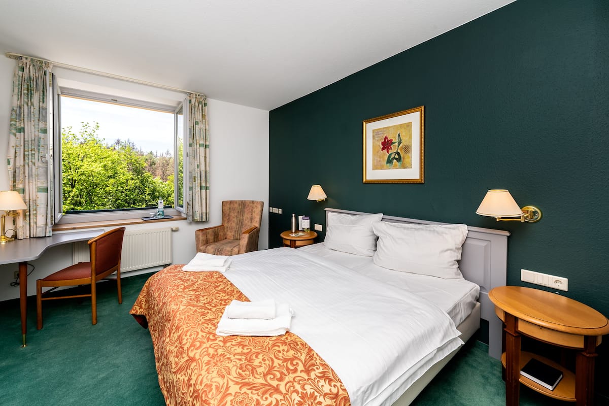 A hotel room with a bed, a desk, a chair, a window with curtains, a nightstand, and a wall-mounted lamp.