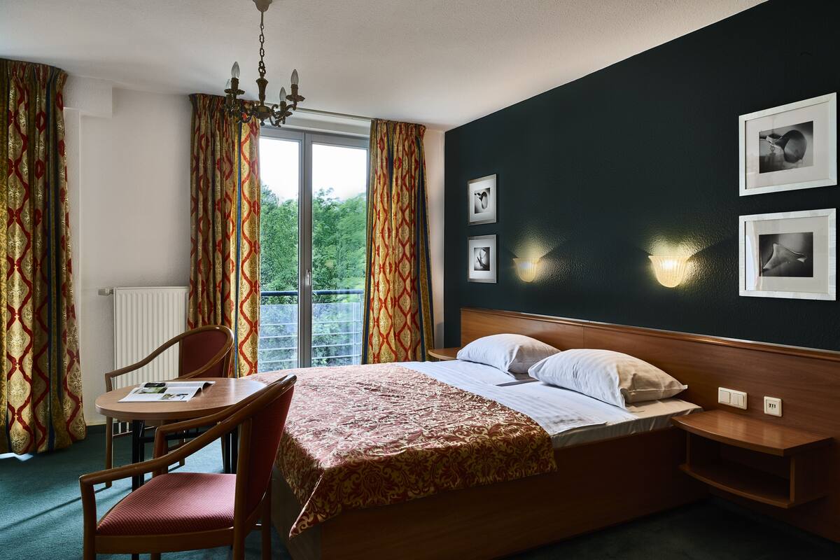 A hotel room with a bed, a desk, a chair, a chandelier, and a view of greenery outside.