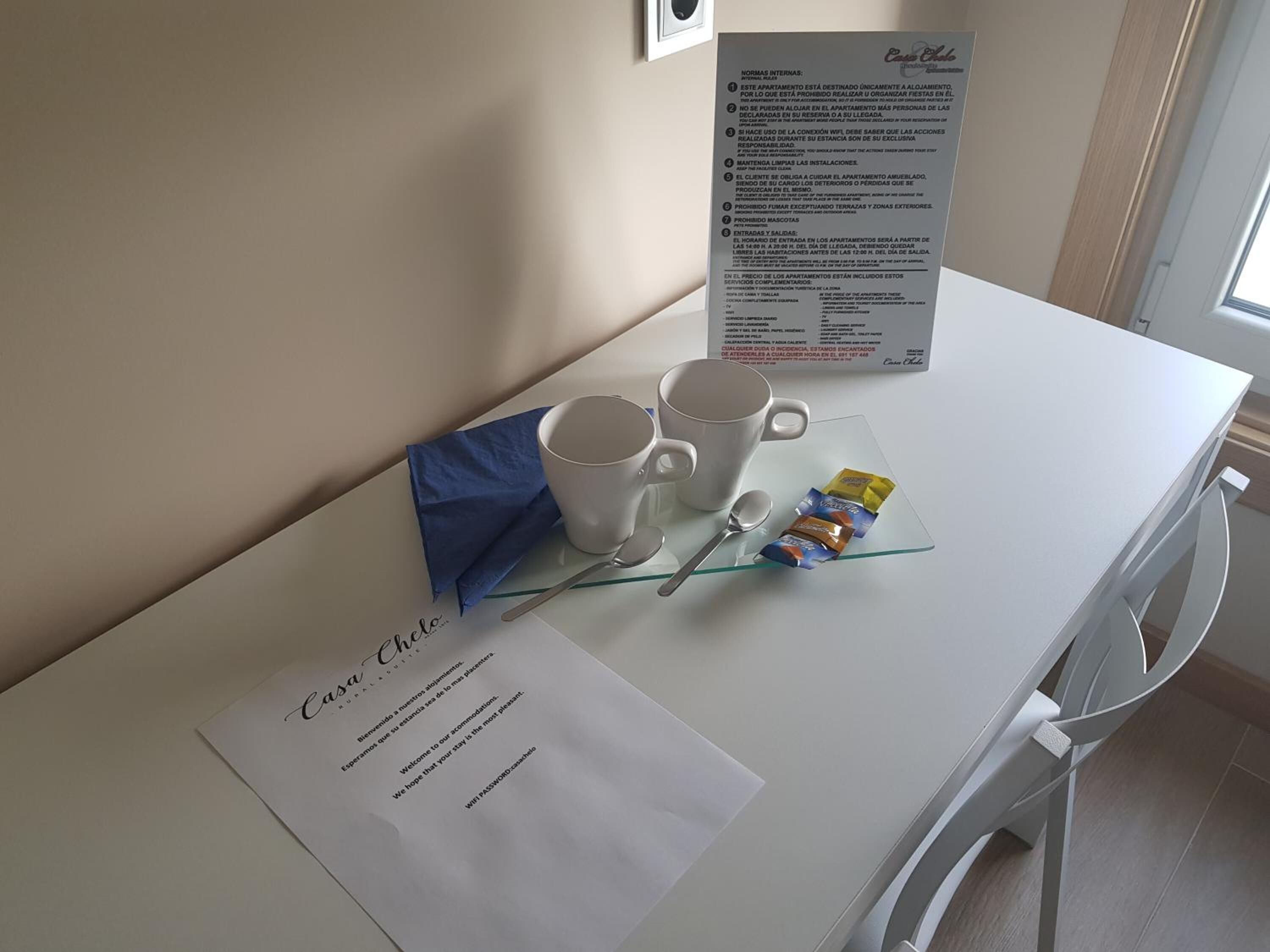 A hotel room with a white table, two white mugs, a spoon, a packet of snacks, and a welcome note.