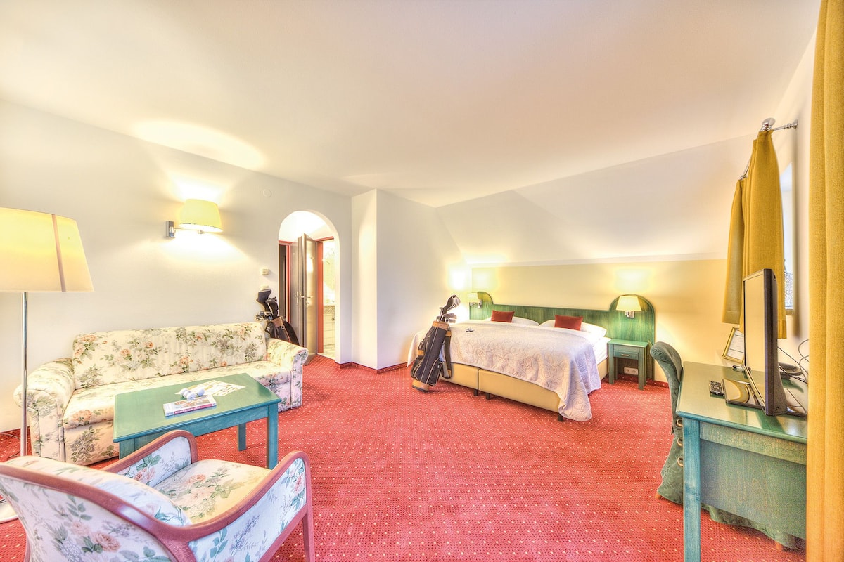 A hotel room with a red carpet, floral sofa, a green armchair, a bed with red pillows, a desk with a lamp, and a mirror.
