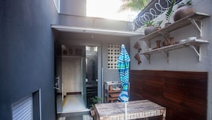 A small outdoor patio area with a wooden table and bench, a striped umbrella, and shelves with decorative items. - Economy Apart (Natal)