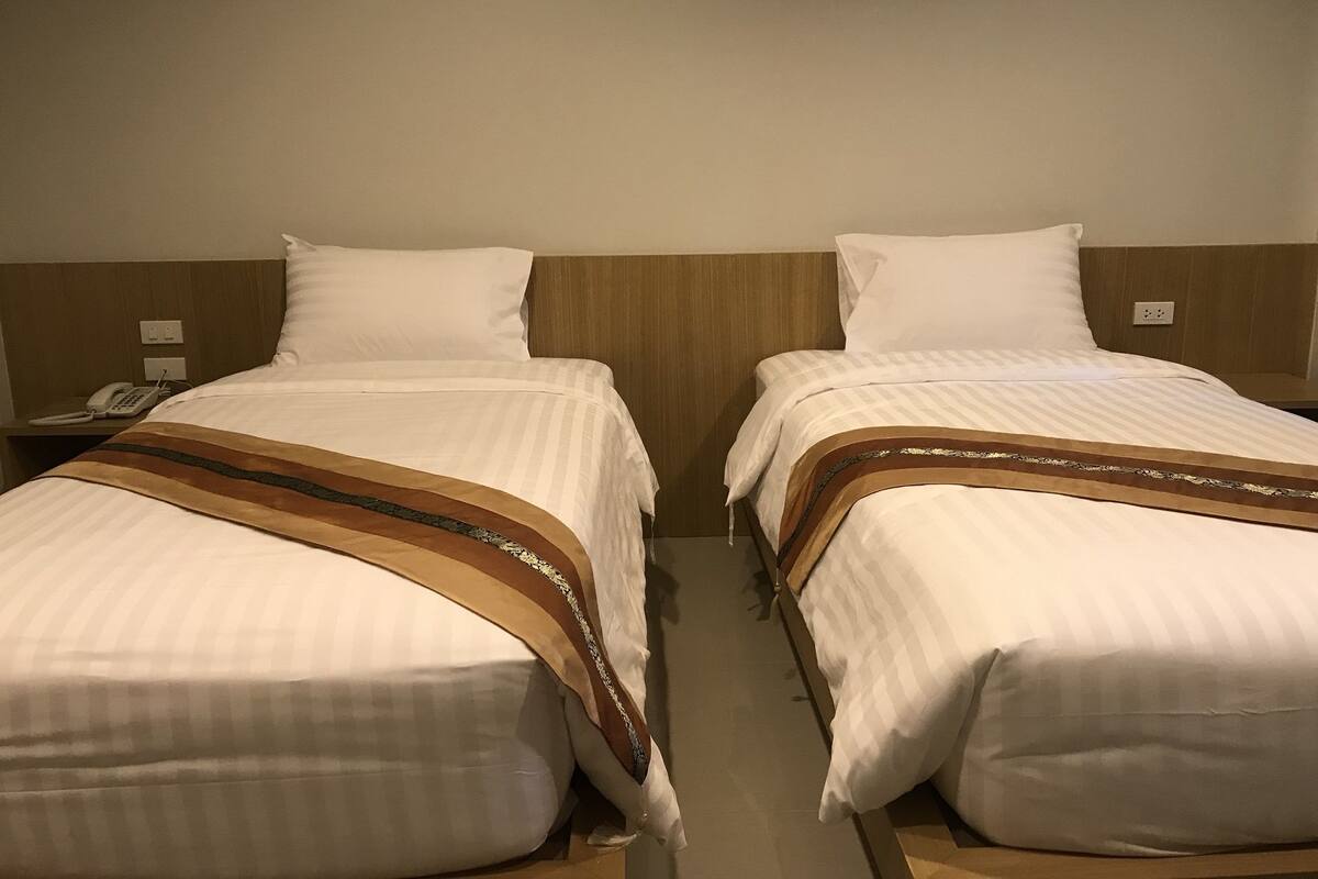 Twee bedden met witte lakens en bruine decoratieve randen, een houten hoofdbord en een telefoon op het nachtkastje.