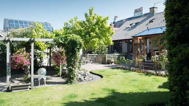 A garden with a pergola, a bench, and a pathway leading to a house with a tiled roof. - Burswood Guest House (York)