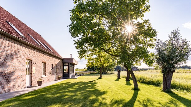 A brick building with a sloped roof, a covered patio, and a well-maintained lawn with trees. - Holiday Home Babillie (Roeselare)