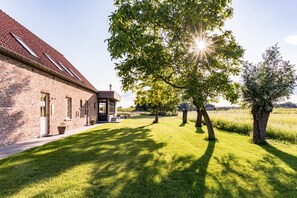 A brick building with a sloped roof, a covered patio, and a well-maintained lawn with trees. - Holiday Home Babillie (Roeselare)