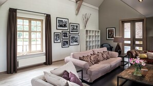 A spacious living room with a high ceiling, a large window with curtains, a beige sofa, a coffee table, and a bookshelf. - Holiday Home Babillie (Roeselare)