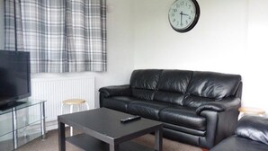 Un salon avec un canapé en cuir noir, une table basse en verre, un téléviseur posé sur un meuble et une horloge au mur.
