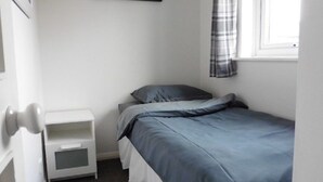 Une petite chambre avec un lit simple, une table de chevet blanche et une fenêtre ornée d’un rideau à carreaux.