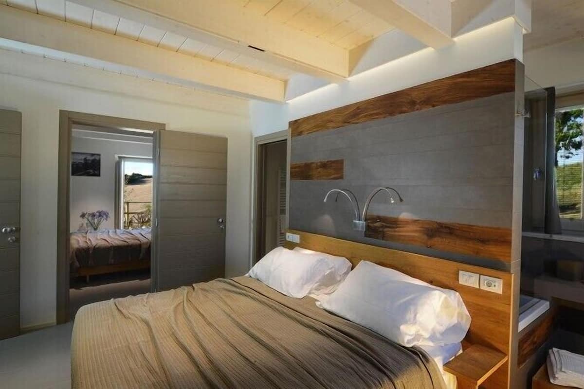 A modern bedroom with a large bed, a wooden headboard, a built-in vanity with a mirror, and a view of the outdoors.