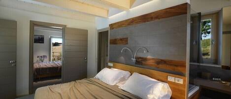 A modern bedroom with a large bed, a wooden headboard, a built-in vanity with a mirror, and a view of the outdoors.